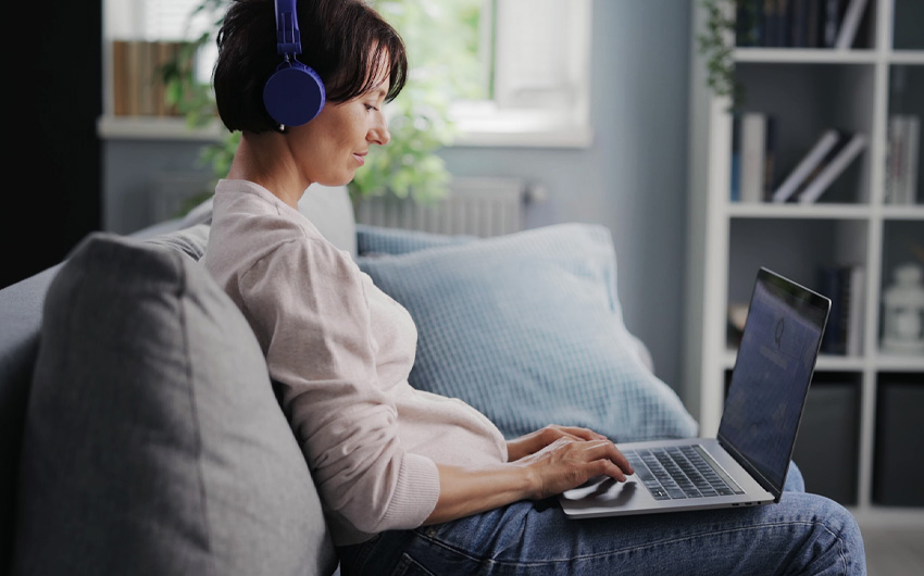 Uma mulher sentada no sofá, com fones de ouvido, utiliza um notebook